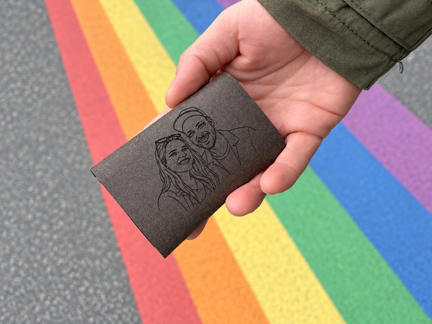 KNIFF Slim in der Hand mit Foto-Gravur: ein Paar als feine Linienzeichnung auf dem braunen LOVR, darunter ein Regenbogen auf dem Asphalt