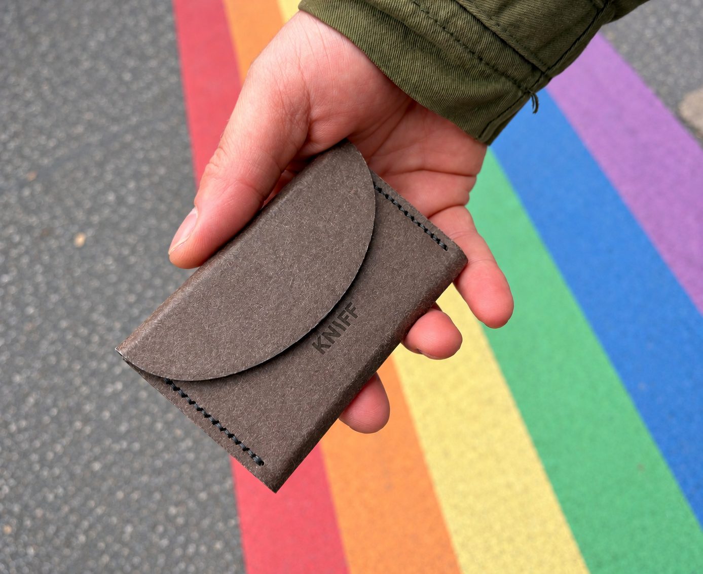 KNIFF Slim in der Hand über einem Regenbogen auf dem Asphalt: runde Klappe, gelasertes KNIFF-Logo, dunkle Sattelnaht